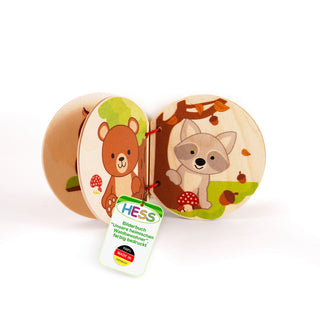„Ein geöffnetes Holz-Bilderbuch für Kinder mit bunten Tierillustrationen. Auf der linken Seite steht ein Bär neben einem Pilz, auf der rechten Seite sitzt ein Fuchs unter einem Baum, umgeben von Eicheln. Ein Etikett am Buch trägt die Aufschrift: ‚HESS Bilderbuch „unsere heimischen Waldbewohner“, farbig bedruckt, 100 % MADE IN GERMANY‘.“