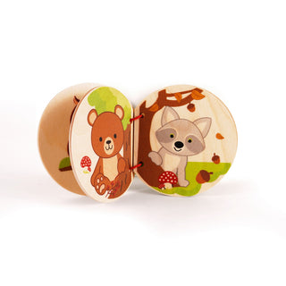 „Ein kleines Holzbuch mit runden Seiten, geöffnet auf zwei farbenfrohe Tierillustrationen in einer Waldszene. Links ist ein Bär mit einer Eichel zu sehen, umgeben von Pilzen und Pflanzen. Rechts steht ein Fuchs neben einem Baum, umgeben von fallenden Blättern und Eicheln.“