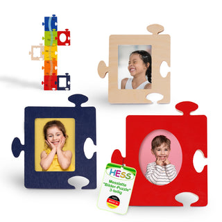 Das Bild zeigt ein Set aus bunten Bilderrahmen in Puzzleteilform und einer Messlatte. Drei große Puzzleteile in Blau, Rot und Naturholz enthalten jeweils ein Kinderfoto. Die Messlatte ist mehrfarbig und mit kleineren Puzzleteilen verziert. Ein Etikett trägt die Aufschrift: „HESS Messlatte 'Bilder-Puzzle' 3-teilig“, hergestellt in Deutschland.