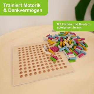 „Ein Holzbrett mit vielen runden Löchern liegt auf einem Tisch. Daneben befindet sich ein Haufen bunter, rechteckiger Stifte in Grün, Blau, Gelb, Rosa und Weiß. Oben links steht in einem grünen Feld: ‚Trainiert Motorik & Denkvermögen‘, oben rechts: ‚Mit Farben und Mustern spielerisch lernen‘.“
