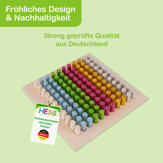 „Ein quadratisches Holzspielzeug mit bunten Steckern in Blau, Grün, Rosa, Gelb und Weiß. Oben links im Bild befindet sich ein grünes Feld mit der Aufschrift: ‚Fröhliches Design & Nachhaltigkeit‘, darunter steht: ‚Streng geprüfte Qualität aus Deutschland‘. Unten links ist ein Etikett mit dem Text: ‚HESS Farbensteckspiel mit hellen Farben 100% MADE IN GERMANY‘.“