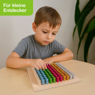 „Ein Kind sitzt an einem Tisch und spielt mit einem Holzspielzeug, das aus mehreren Reihen farbiger Stifte besteht. Im Hintergrund steht eine grüne Pflanze. Oben links im Bild befindet sich ein grünes Schild mit weißer Schrift: ‚Für kleine Entdecker‘.“