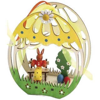 Farbiges Fensterbild aus Holz in 3D Optik. Das Festerbild ist grün und gelb bemalt. Im Bild befindet sich eine farbige Osterhäsin auf der Bank mit Ostereiern und einem Bäumchen.