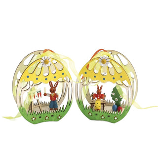 2 Fensterbilder aus Holz in 3D-Optik. Die Fensterbilder sind grün und gelb bemalt. In einem Bild befindet sich ein farbiger Osterhasenlehrer mit einer Schultafel und im zweiten Bild eine farbige Osterhäsin auf der Bank mit Ostereiern und einem Bäumchen