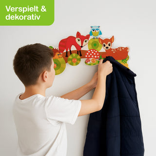 „Ein Junge hängt eine dunkelblaue Jacke an eine bunte Garderobe mit Waldtieren. Zu sehen sind Illustrationen eines Fuchses, einer Eule, eines Rehs und eines Igels sowie Pilze und grüne Kreise als Dekoration. Oben links steht ‚Verspielt & dekorativ‘.“