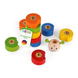 Ein buntes Holzspielzeug in Form einer Raupe, bestehend aus mehreren runden Holzstücken in Grün, Rot, Blau, Gelb und Orange. Jedes Stück hat eine metallene Öffnung in der Mitte. Auf einem grünen Segment befindet sich ein Etikett mit der Aufschrift „HESS Geburtstagsleuchter 'Raupe' 100% MADE IN GERMANY“. Daneben liegt ein Holzkopf mit einem freundlichen Gesicht und zwei grünen Ohren oder Fühlern.