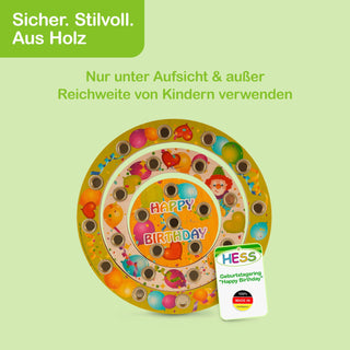 Runder Geburtstagsring aus Holz mit bunten Motiven wie Luftballons, Herzen und Figuren. In der Mitte steht „HAPPY BIRTHDAY“. Der Ring enthält mehrere Löcher für Kerzen oder Dekorationen. Oben links im Bild befindet sich ein grünes Feld mit dem Hinweis „Sicher. Stilvoll. Aus Holz“ und einem Sicherheitstext. Unten rechts ist eine Verpackung mit dem Aufdruck „HESS Geburtstagsring 'Happy Birthday' 100% MADE IN GERMANY“ sichtbar.