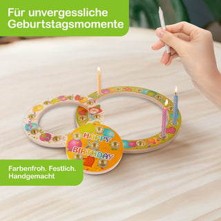 Eine Hand steckt eine brennende Geburtstagskerze in einen dekorativen Kerzenhalter aus zwei ineinander verschlungenen, bunt bemalten Holzringen. Auf einem der Ringe steht „HAPPY BIRTHDAY“. Mehrere Kerzen sind bereits entzündet. Oben links im Bild steht „Für unvergessliche Geburtstagsmomente“, unten links „Farbenfroh. Festlich. Handgemacht“.