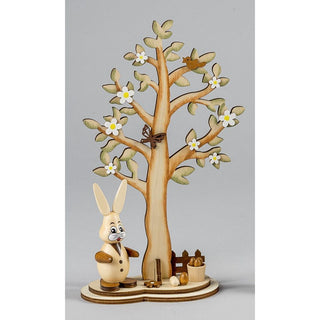 Dekorative Holzszene mit einem aufrecht stehenden Hasen in Weste und Knöpfen im Vordergrund. Dahinter ein großer Baum mit grünen Blättern und weißen Blüten. Auf einem Ast sitzt ein Vogel, in der Mitte des Baumes ein Schmetterling. Am Baumfuß liegen bunte Eier und ein kleiner Holzeimer.