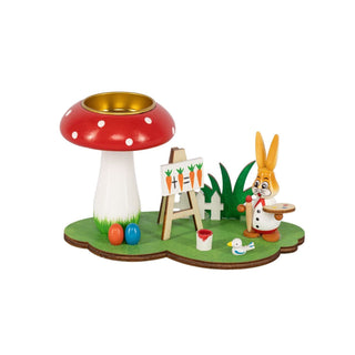 Grünfarbener Sockel mit einem Fliegenpilz als Teelichthalter und einem farbigen Osterhasen als Maler und einer Staffelei