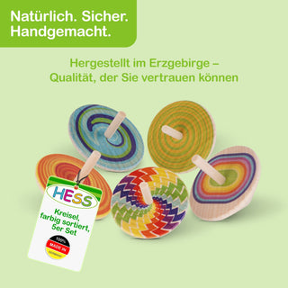 Fünf bunte Holzkreisel mit unterschiedlichen Mustern und Farben liegen auf einem hellgrünen Hintergrund. Oben links steht in weißer Schrift „Natürlich. Sicher. Handgemacht.“ und darunter in Grün „Hergestellt im Erzgebirge – Qualität, der Sie vertrauen können“. Unten links ist ein Etikett mit dem Schriftzug „HESS – Kreisel, farbig sortiert, 5er Set“ sowie einem roten Siegel mit „100% MADE IN GERMANY“.