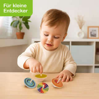 Ein Kleinkind sitzt an einem Tisch und spielt mit vier bunten Holzkreiseln. Im Hintergrund sind ein Regal mit Dekoration und eine Pflanze auf einem Fensterbrett zu sehen. Oben links im Bild steht in weißer Schrift auf grünem Hintergrund: „Für kleine Entdecker“.