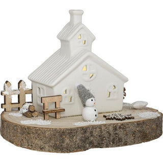 Weißes LED-Keramikhaus mit warmem Licht auf Holzsockel, 16 cm hoch, dekoriert mit Schneemann in grauer Mütze, Holzbank, Holzscheiten, Zaun und Kunstschnee – winterliches Arrangement mit Weihnachtsflair.