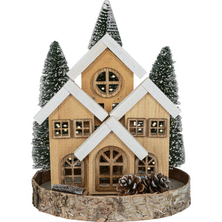 LED-Dekoration in Form eines winterlichen Waldhauses auf einem naturbelassenen Holzsockel, umgeben von drei schneebedeckten Tannenbäumen. Das Haus ist mit warm leuchtenden LED-Lichtern ausgestattet, die durch Fenster und Türen scheinen. Kleine Deko-Elemente wie Tannenzapfen und Holzstücke verzieren den Sockel. Die Höhe beträgt 24 cm.