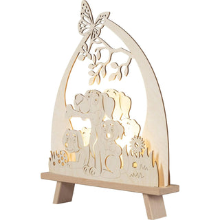 Die LED Standleuchte "Hundefreund" aus Holz, zeigt drei kleine Fellnasen, die fröhlich im Gras sitzen. Einer der Hunde hält stolz einen Knochen im Maul, während auf einem Ast von einem Baum ein zarter Schmetterling Platz genommen hat. Beleuchtete Schrägansicht.