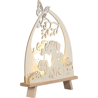 Die LED Standleuchte "Hundefreund" aus Holz, zeigt drei kleine Fellnasen, die fröhlich im Gras sitzen. Einer der Hunde hält stolz einen Knochen im Maul, während auf einem Ast von einem Baum ein zarter Schmetterling Platz genommen hat. Beleuchtete Schrägansicht.