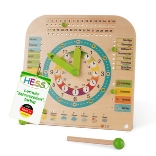 Farbig gestaltete Lernuhr aus Holz mit grünen Zeigern. Links sind die Monate, rechts die Wochentage und Jahreszeiten dargestellt. Unten befinden sich Symbole für Wetterbedingungen. Ein Schild zeigt: „HESS Lernuhr 'Jahreszeiten', farbig 100% MADE IN GERMANY“.