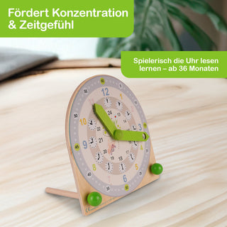 Holz-Lernuhr mit großen grünen Zeigern, Zahlen von 1–12 und 13–24 sowie Minutenmarkierungen in 5er-Schritten. Die Uhr steht auf einem Tisch, im Hintergrund ist ein unscharfes Pflanzenblatt zu sehen. Text im Bild: „Fördert Konzentration & Zeitgefühl“ und „Spielerisch die Uhr lesen lernen – ab 36 Monaten“.