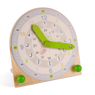 Holz-Lernuhr mit zwei grünen Zeigern und drei konzentrischen Kreisen: außen Minuten in 5er-Schritten (0–60), mittig Stunden von 1–12 in bunten Farben, innen Stunden von 13–24. In der Mitte sind kleine Uhren mit Stundenangaben sowie ein Schmetterling und eine Blume abgebildet.
