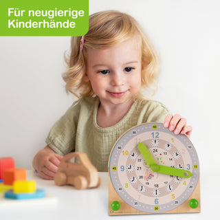 Ein kleines Kind sitzt an einem Tisch mit einer Holzspieluhr und weiteren bunten Spielzeugen. Die Uhr hat grüne Zeiger und zeigt Zahlen in Blau, Gelb und Schwarz. Oben links steht auf grünem Hintergrund der Text „Für neugierige Kinderhände“.