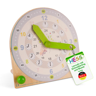 Holz-Lernuhr mit zwei grünen Zeigern und doppelter Ziffernanzeige: außen Stunden von 1–12 in bunten Farben, innen Stunden von 13–24 in Schwarz. In der Mitte sind ein Schmetterling und eine Blume abgebildet. Kleine Uhren zeigen jeweils eine Stunde. Ein Etikett trägt die Aufschrift „HESS Lernuhr mit Schmetterling und Blume farbig“ und „100% MADE IN GERMANY“.