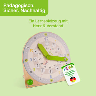 Runde Holz-Lernuhr mit zwei grünen Zeigern. Die äußere Skala zeigt die Stunden 1–12 in Blau und Gelb, die innere Skala die Stunden 13–24 in Grau. Auf dem Zifferblatt sind kleine Tiere und Pflanzen abgebildet. Oben links steht „Pädagogisch. Sicher. Nachhaltig“, darunter „Ein Lernspielzeug mit Herz & Verstand“. Ein Etikett an der Uhr trägt den Text „HESS Lernuhr mit Schmetterling und Blume“ sowie ein rotes Siegel „Made in Germany“.