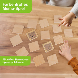 Zwei Kinderhände spielen ein Memo-Spiel auf einem Holztisch. Die quadratischen Karten sind größtenteils umgedreht, zwei zeigen ein Bärenmotiv. Der Text „Farbenfrohes Memo-Spiel“ und „Mit süßen Tiermotiven spielerisch lernen“ ist in grüner Schrift in den Ecken des Bildes zu sehen.