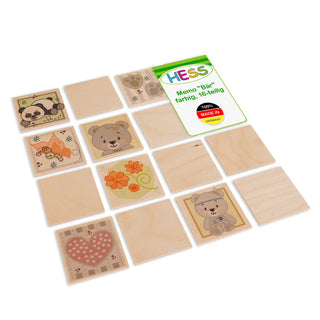 Holz-Memo-Spiel mit 16 quadratischen Plättchen in einer 4×4-Anordnung. Einige Plättchen zeigen bunte Motive wie einen Panda, einen Bären, eine orangefarbene Blume und ein Herz mit Punkten. Die Verpackung ist sichtbar und trägt die Aufschrift „HESS Memo 'Bär' farbig, 16-teilig 100% MADE IN GERMANY“.