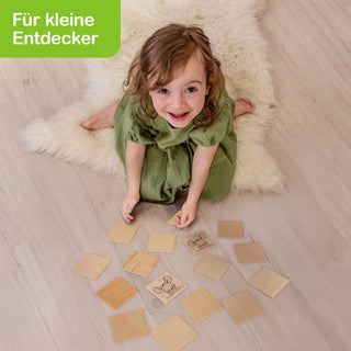 Ein Kind sitzt auf einem flauschigen Teppich und spielt mit einem Memo-Spiel aus quadratischen Holzplättchen. Zwei Plättchen zeigen eine Giraffe und eine Maus, andere sind umgedreht. Oben links steht „Für kleine Entdecker“ auf grünem Hintergrund.