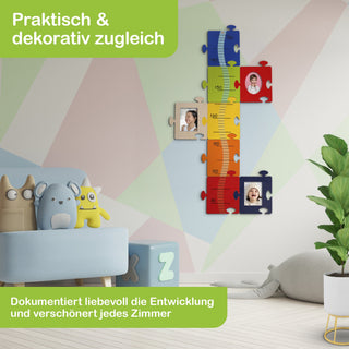 Kinderzimmer mit bunter Messlatte in Puzzleform an der Wand, mit Höhenangaben von 70 cm bis 150 cm und Platz für Fotos. Auf einem hellblauen Sofa liegen Stofftiere, auf dem Boden befinden sich Alphabetbausteine. Text im Bild: „Praktisch & dekorativ zugleich“ und „Dokumentiert liebevoll die Entwicklung und verschönert jedes Zimmer.“