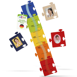 Farbiges Puzzle in Form einer Messlatte mit Höhenangaben von 90 cm (Rot) bis 150 cm (Blau). Die Messlatte ist in fünf farbige Abschnitte unterteilt: Rot, Orange, Gelb, Grün und Blau. Links befindet sich ein Schild mit der Aufschrift „HESS Messlatte 'Bilder-Puzzle' farbig“ und „100% Made in Germany“. Um die Messlatte sind vier Puzzleteile mit Bildern von Personen angeordnet.
