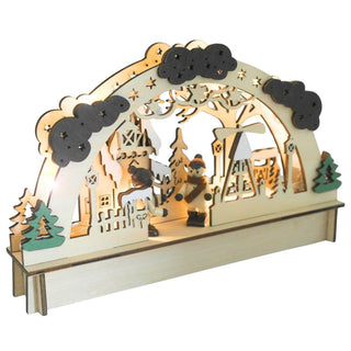 Beleuchteter LED-Holz-Lichterbogen mit filigranen Ausschnitten zeigt eine winterliche Dorfszene: Zwei kleine Figuren in Winterkleidung stehen im Zentrum, umgeben von Bäumen, einer Pyramide und Gebäuden. Der obere Bogen ist mit Wolken und Sternen verziert. Links und rechts am Sockel befinden sich grüne Bäume. Schrägansicht