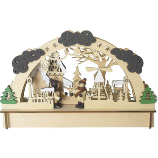 LED-Holz-Lichterbogen mit filigranen Ausschnitten zeigt eine winterliche Dorfszene: Zwei kleine Figuren in Winterkleidung stehen im Zentrum, umgeben von Bäumen, einer Pyramide und Gebäuden. Der obere Bogen ist mit Wolken und Sternen verziert. Links und rechts am Sockel befinden sich grüne Bäume. 