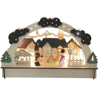 Beleuchteter LED-Holz-Lichterbogen mit einem liebevoll gestalteten Dorfmotiv: Mehrere Häuser mit detaillierten Fenstern und Dächern sowie eine Kirche mit Turm stehen im Zentrum. Links und rechts rahmen zwei Bäume die Szene ein. Im Vordergrund befinden sich eine Holzfigur in Winterkleidung und ein Schneemann mit rotem Schal und schwarzem Hut. Ein Bogen mit Sternen und Wolken überspannt das gesamte Arrangement.