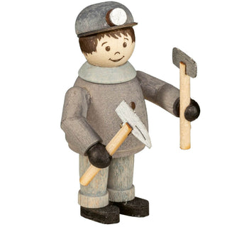 **Alt Text:** Die Abbildung zeigt eine Holzfigur eines Bergmanns. Der Bergmann trägt eine graue Kleidung mit schwarzen Handschuhen und Stiefeln sowie einen grauen Helm mit einer Grubenlampe vorne. Die Figur hält zwei Werkzeuge: eine Axt in der linken Hand und einen Hammer in der rechten Hand. Der Bergmann hat kurzes braunes Haar und einfache, bemalte Gesichtszüge.