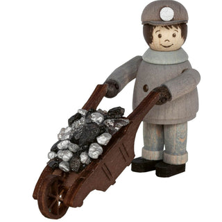 Die Abbildung zeigt eine Holzfigur einer Person in grauer Kleidung und Mütze, die eine braune Schubkarre mit Erz und Silber schiebt. Die Figur hat einfache, bemalte Gesichtszüge, darunter Augen, Augenbrauen und ein Lächeln. Die Schubkarre hat zwei Griffe und ein Rad vorne.