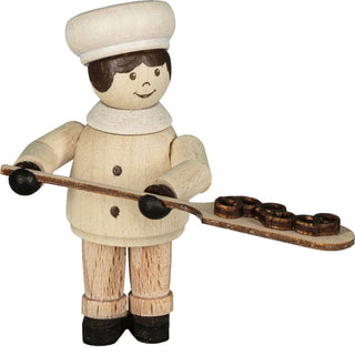 "Eine Holzfigur zeigt einen Bäcker, der ein langes Holzbrett mit drei Brezeln darauf hält. Die Figur trägt eine weiße Bäckermütze und einen weißen Mantel sowie schwarze Handschuhe und Schuhe. Das Gesicht der Figur ist einfach bemalt, einschließlich Augen, Augenbrauen und einem lächelnden Mund."