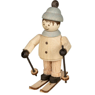 Die Abbildung zeigt eine Holzfigur einer Person beim Skifahren. Die Figur ist in Winterkleidung gekleidet, einschließlich einer Mütze mit einem Bommel oben, und hält Skistöcke in beiden Händen. Die Skier sind an den Füßen der Figur befestigt.