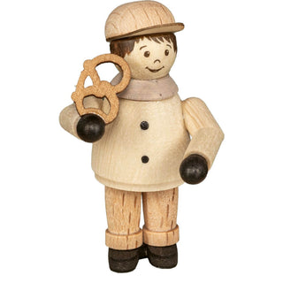 Die Abbildung zeigt eine Holzfigur einer Person, die einen Hut trägt und eine Brezel hält. Die Figur hat einfache Gesichtszüge, mit zwei Punkten für die Augen und einer gebogenen Linie für den Mund. Die Person ist in heller Kleidung mit dunklen Schuhen und Handschuhen gekleidet. 