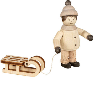 "Eine Holzfigur zeigt eine Person in Winterkleidung, einschließlich einer Mütze und einem Mantel, die einen kleinen Holzschlitten an einer Schnur zieht. Die Figur hat einfache Gesichtszüge und steht aufrecht. Der Schlitten ist mit zwei Kufen und Latten oben gestaltet."