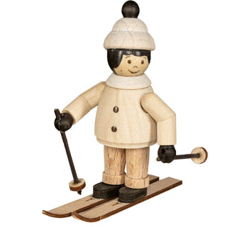 "Eine Holzfigur zeigt eine Person in Winterkleidung, einschließlich einer weißen Mütze mit einem schwarzen Pompon, einem weißen Mantel mit schwarzen Knöpfen und schwarzen Handschuhen. Die Figur steht auf Holzskiern und hält Skistöcke. Das Gesicht der Figur ist einfach und lächelnd bemalt."