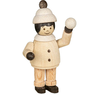 Eine Holzfigur zeigt eine Person in Winterkleidung. Die Figur trägt einen beigen Mantel mit zwei Knöpfen, eine beige Hose und schwarze Schuhe. Die Person hat eine beige Mütze mit einem Bommel und schwarze Haare. Die Figur hält einen Schneeball in der rechten Hand.