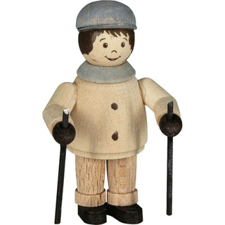 "Eine Holzfigur zeigt eine Person in Winterkleidung. Die Figur trägt einen hellbraunen Mantel, eine beige Hose, schwarze Schuhe und eine graue Mütze. Die Figur hält Nordic Walking Stöcke in der Hand. Das Gesicht der Figur ist mit einfachen Merkmalen wie Augen, Augenbrauen und einem lächelnden Mund bemalt."