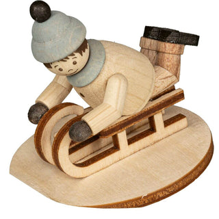 "Eine Holzfigur zeigt eine Person, die auf einem Schlitten liegt. Die Figur trägt eine graue Mütze mit einem schwarzen Bommel sowie schwarze Handschuhe und Stiefel. Der Schlitten steht auf einer ovalen Holzplatte."