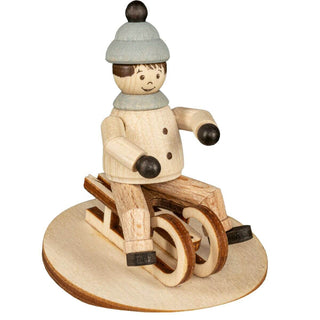 "Eine Holzfigur zeigt eine Person, die eine hellblaue Mütze und einen beigen Mantel mit schwarzen Knöpfen trägt und auf einem Holzschlitten sitzt. Die Figur ist auf einer ovalen Holzplatte montiert. Die Person hat schwarze Handschuhe und Stiefel, und der Schlitten hat zwei Kufen."