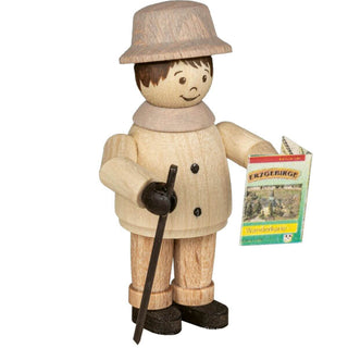 **Alt Text:** Die Abbildung zeigt eine Holzfigur einer Person, die einen Hut trägt und in einer Hand einen Wanderstock sowie in der anderen ein Heft hält. Auf dem Heft steht der Text "ERZGEBIRGE" zusammen mit einer Landschaftsillustration. Die Figur hat einfache Gesichtszüge mit gemalten Augen, Augenbrauen und Mund. Die Person ist in heller Kleidung und dunklen Schuhen gekleidet.