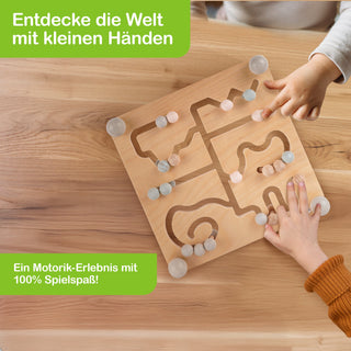 Zwei Kinderhände spielen mit einem hölzernen Labyrinth-Spiel auf einem Holztisch. Das Spielbrett enthält verschlungene Pfade, in denen bunte Kugeln bewegt werden können. Oben links steht in weißer Schrift auf grünem Hintergrund: ‚Entdecke die Welt mit kleinen Händen‘. Unten links steht: ‚Ein Motorik-Erlebnis mit 100% Spielspaß!