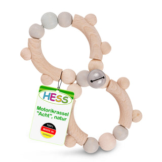Holzrassel in Form einer Acht, bestehend aus naturbelassenen Holzteilen und hell lackierten Holzperlen. An einem Abschnitt ist eine kleine silberne Glocke befestigt. Ein Etikett am Spielzeug trägt die Aufschrift: ‚HESS Motorikrassel ‚Acht‘, natur 100% MADE IN GERMANY‘.