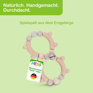 Holzspielzeug in Form einer Acht, bezeichnet als Motorikrassel, bestehend aus naturfarbenen Holzkugeln und -stäben, die durch elastische Bänder verbunden sind. Eine kleine Glocke ist integriert. Oben links im Bild steht in einem grünen Feld: ‚Natürlich. Handgemacht. Durchdacht.‘ Darunter: ‚Spielspaß aus dem Erzgebirge‘. Ein Etikett am Spielzeug trägt die Aufschrift ‚HESS Motorikrassel ‚Acht‘, natur‘ sowie ‚100% MADE IN GERMANY‘.