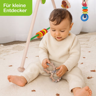 Ein kleines Kind sitzt auf einem hellen Teppich und spielt mit einem grauen Beißring. Im Hintergrund steht ein Holzspielbogen mit bunten Ringen und Spielzeugen. Oben links im Bild befindet sich ein grünes Textfeld mit der weißen Aufschrift ‚Für kleine Entdecker‘.